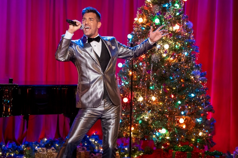 A person singing into a microphone on a stage with a christmas treeDescription automatically generated with medium confidence